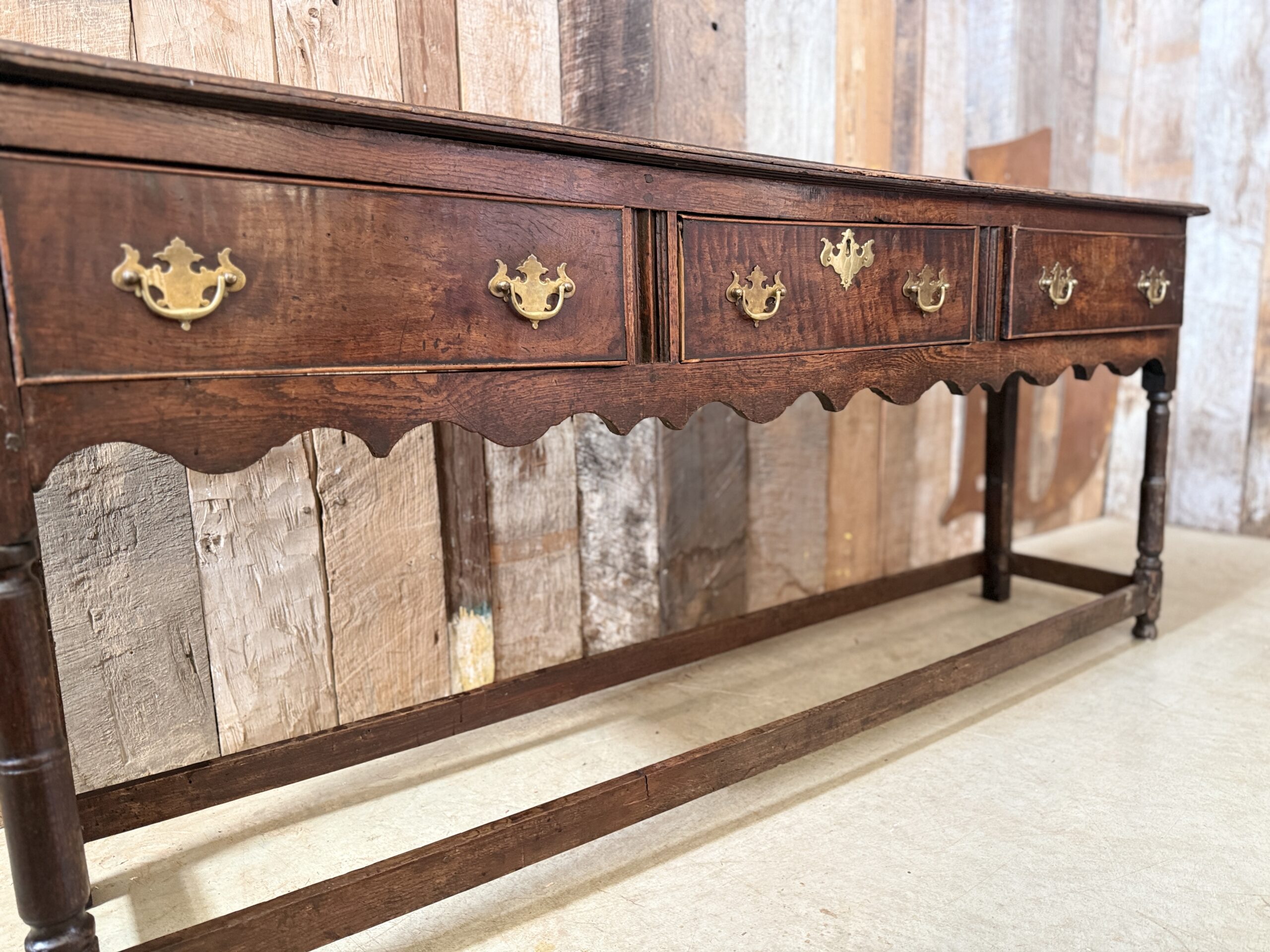 Antique George III English oak dresser base circa 1780 with three drawers, brass swan-neck handles, shaped apron and turned legs.